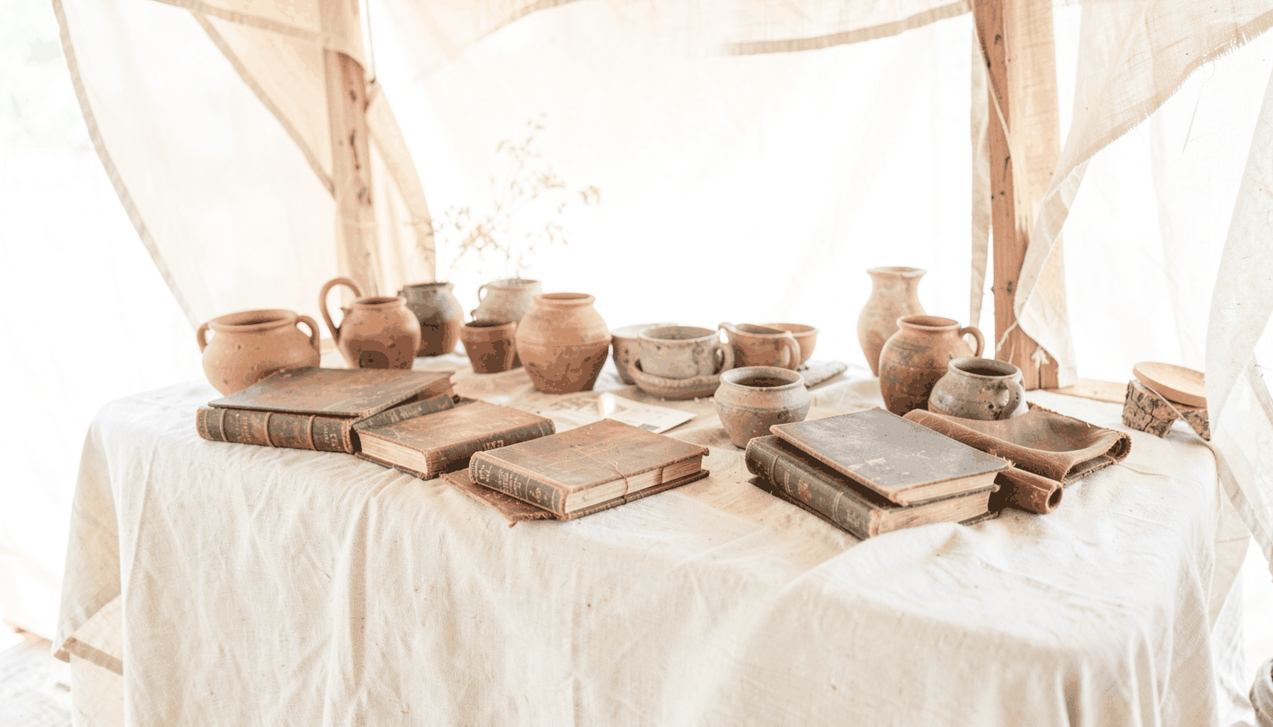A bright, airy photograph of a rustic wooden market stall draped in natural linen, with handcrafted pottery and leather-bound books arranged across its surface, bathed in soft daylight.