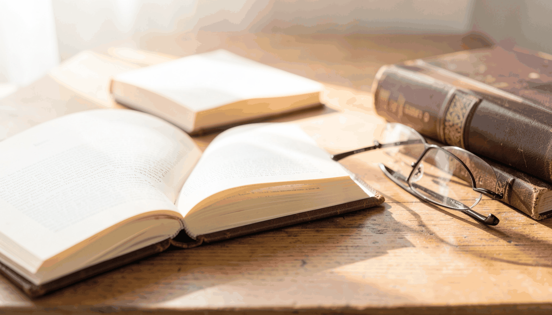 A sunlit wooden table with several open hardback books, their cream pages catching warm light; a pair of reading glasses rests beside a leather-bound volume, with soft shadows and a calm, scholarly atmosphere